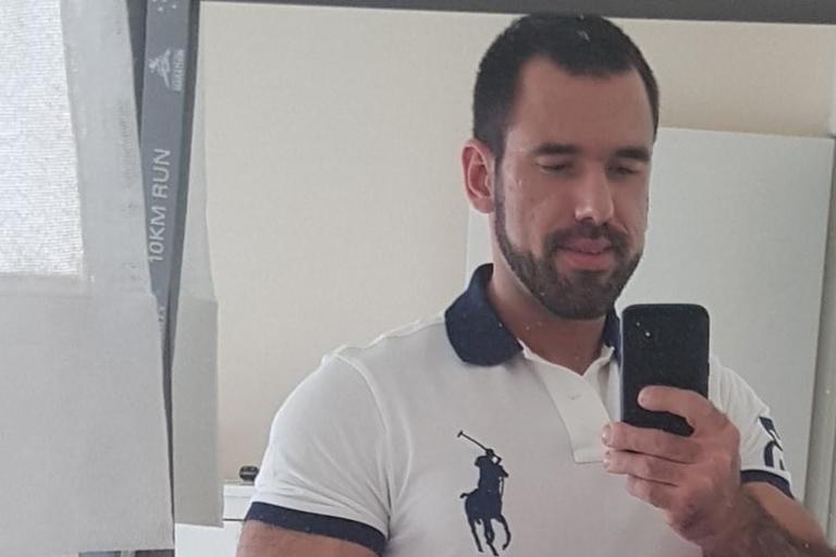 A man with dark hair and facial hair wears a polo shirt and smiles in a mirror selfie