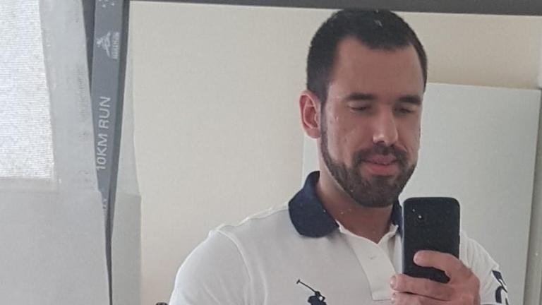 A man with dark hair and facial hair wears a polo shirt and smiles in a mirror selfie
