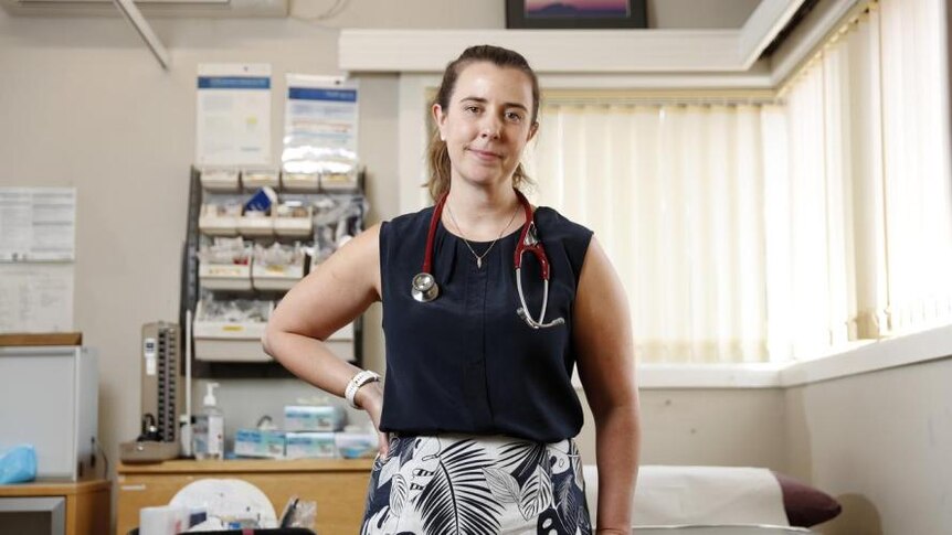 Woman stands in medical room with stethoscope around her neck. 