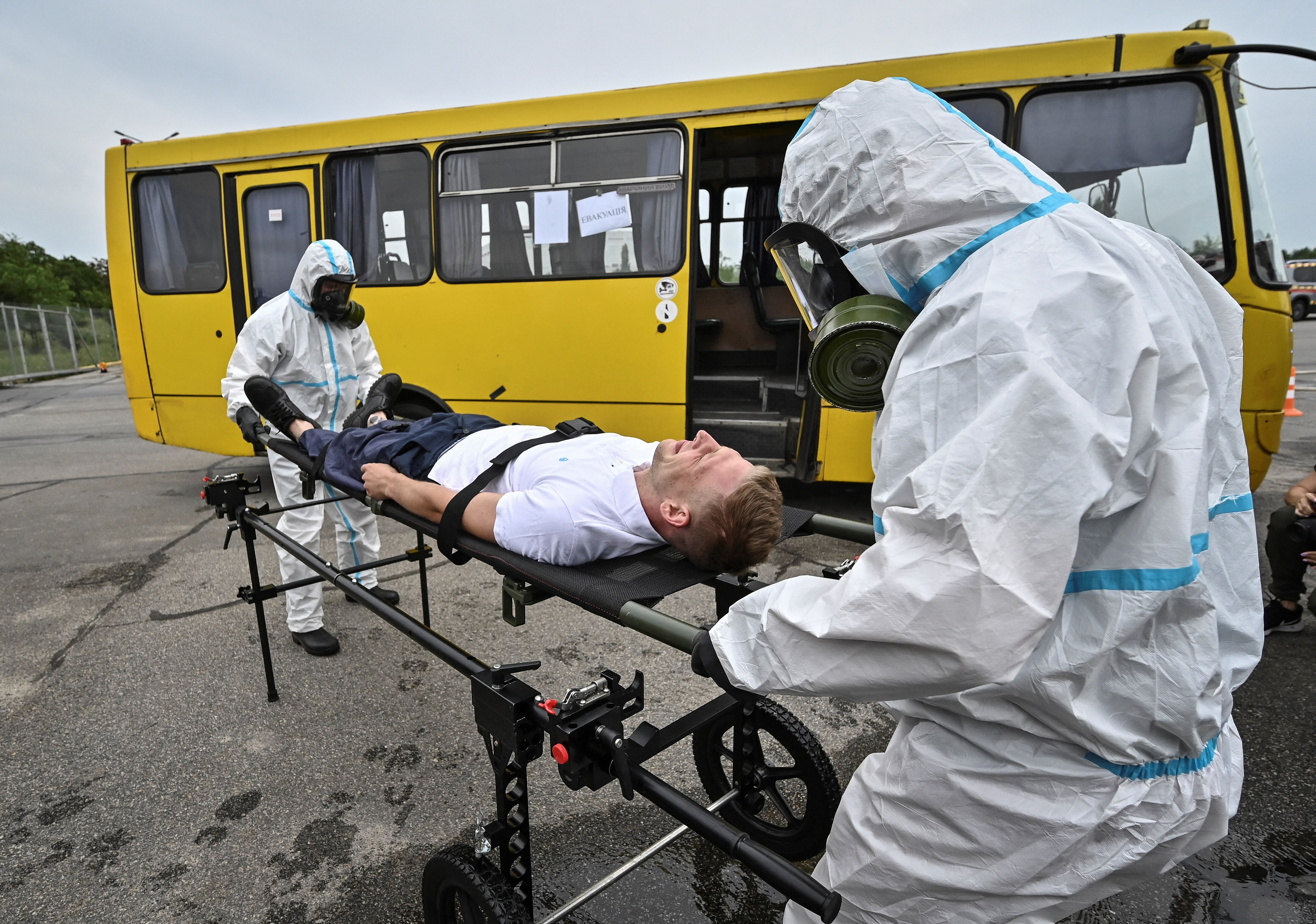 Two men in white hazmat suits wheel another man in civilian clothes strapped to a gurney past a big yellow bus.