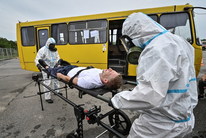 Two men in white hazmat suits wheel another man in civilian clothes strapped to a gurney past a big yellow bus.