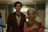 Middle-aged man and woman dressed in 50s attire walk through elegantly decorated house foyer. Woman carries baby in arms.