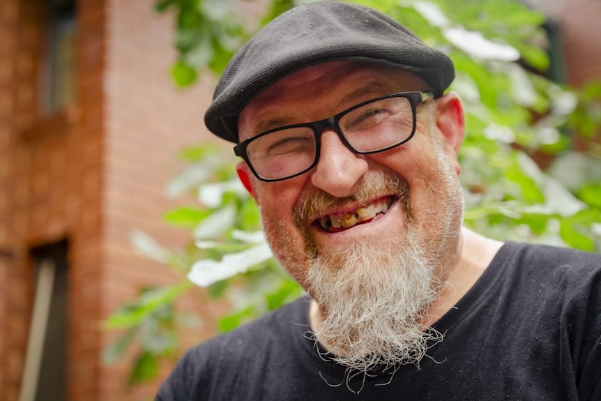 A man with a beard wears a hat and smiles, with bushes in the background.