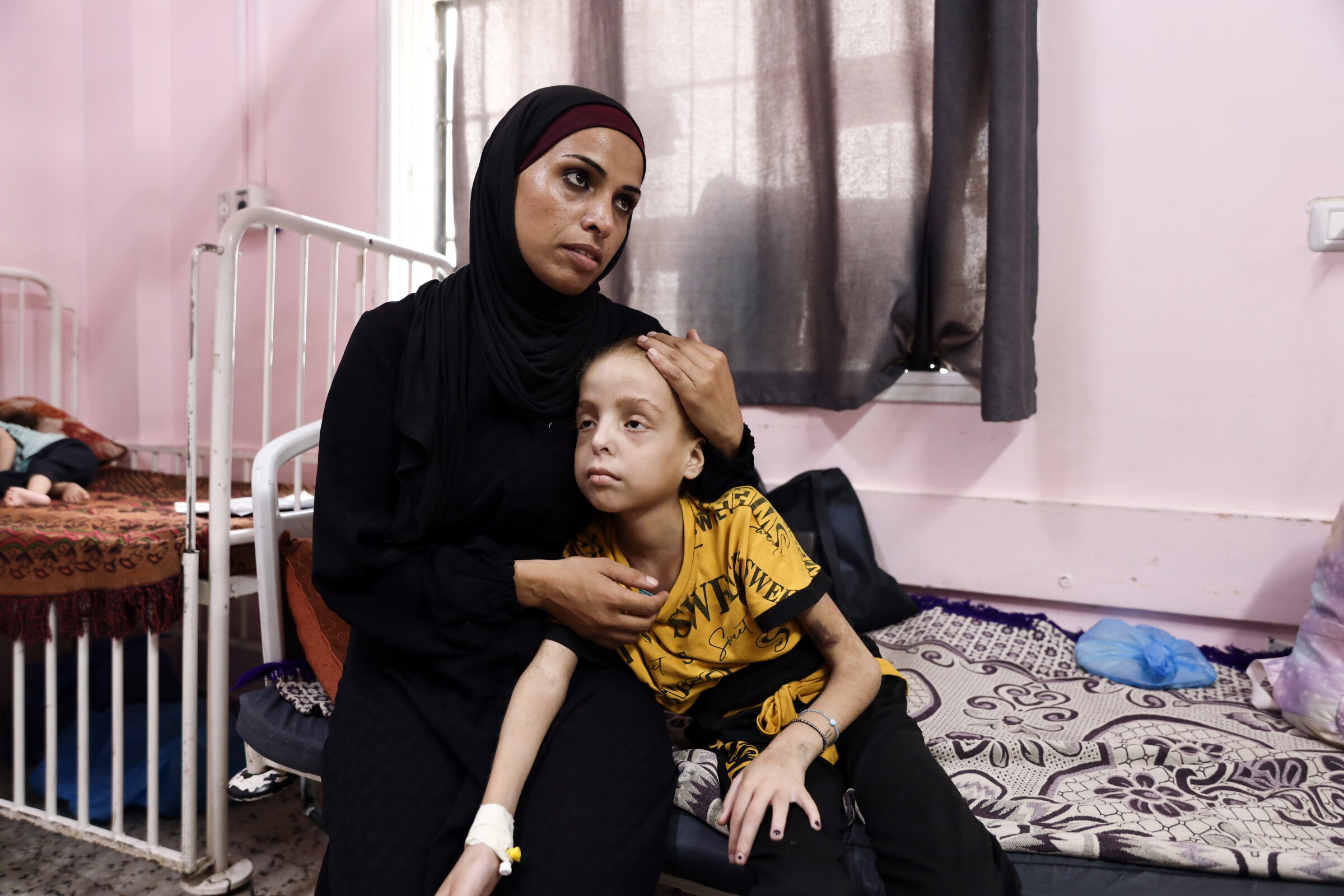A woman in a black hijab holds her daughter, a young girl who looks thin and pale, while sitting in hospital.