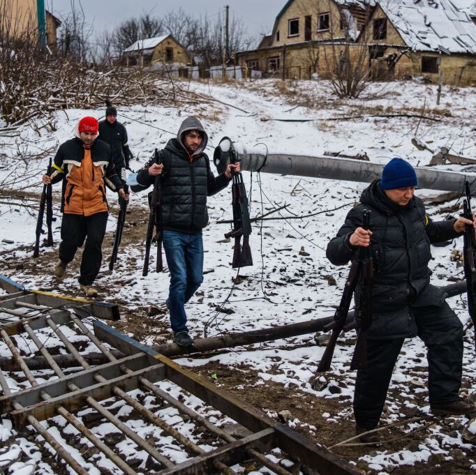 Ukrainian men in casual winter clothes carrying multiple rifles walk along broken bridge in the snow