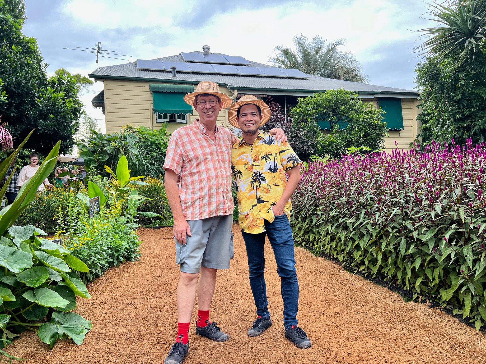 Jerry Coleby-Williams stands with his arm around partner Thuan, both smiling in a bright garden in front of a house