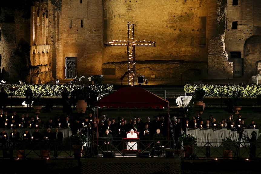 A cross  lights up in front of the Colosseum in Rome with large crowds in front