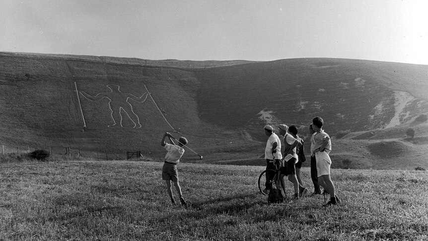 Wide black and white archive shot of men playing golf in a field with an enormous man carved into the hill beyond.