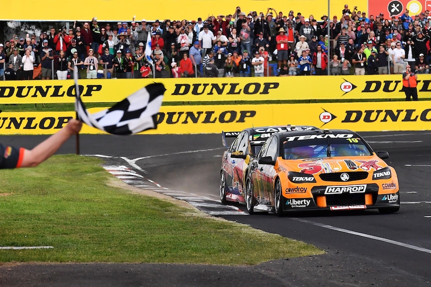 An official holds a chequered flag as two cars approach the finish line in the Bathurst 1000,m with the number 19 car in front.
