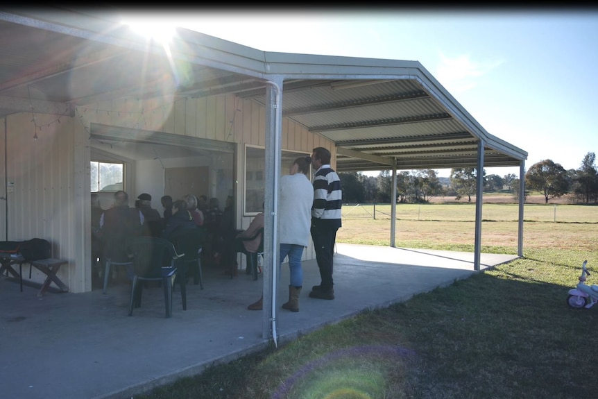 A meeting. People sitting in a shed, two people stand at the back.