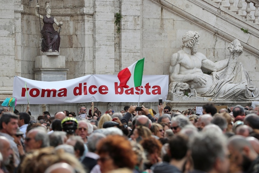 Hundreds of people pack a public square in Rome. Some hold a banner with Italian text that translates to: "Rome says enough".