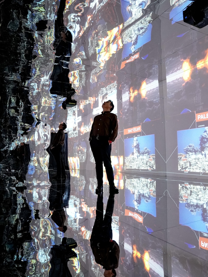 A man stands inside a mirrored room with art projected onto the walls, looking up at the ceiling.