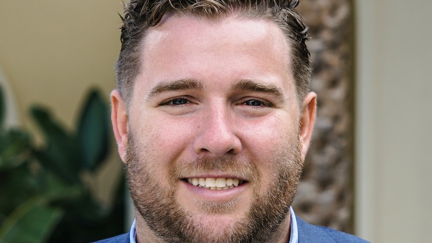 A close-up of a man in blue suit jacket and shirt, with short facial hair, smiling widely.