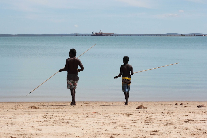 A young man and an older man with traditional Indigenous fishing spears walking into an ocean.