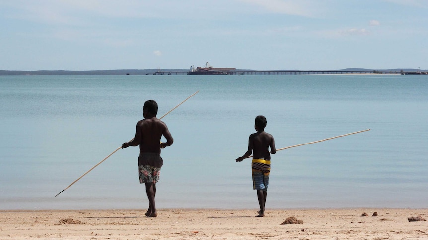 A young man and an older man with traditional Indigenous fishing spears walking into an ocean.