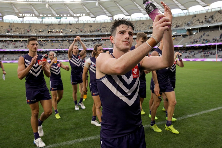 Holding a bottle, Andrew Brayshaw applauds and looks up at the crowd. His teammates are behind him