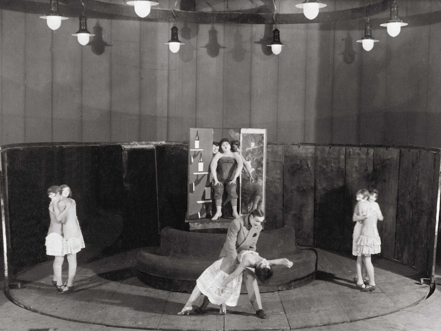Black and white image of a stage with performers including women embracing, and a man and woman dancing at centre.