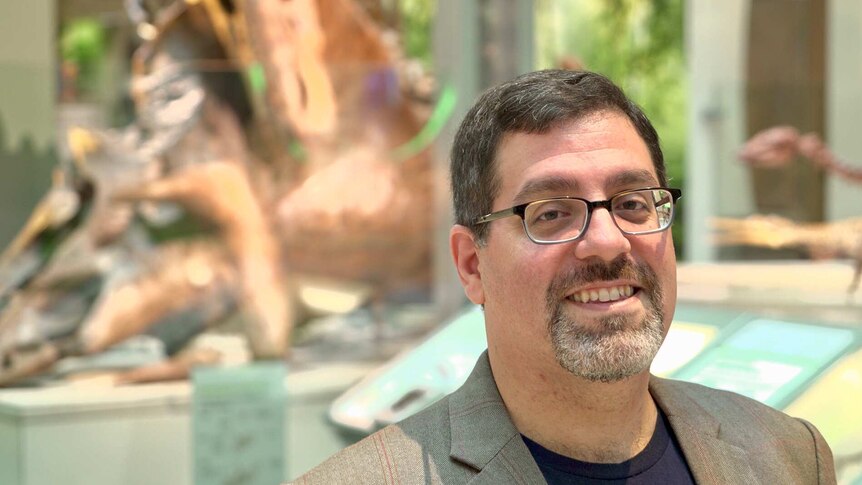 A man in glasses smiles while standing in front of dinosaur fossils at a museum