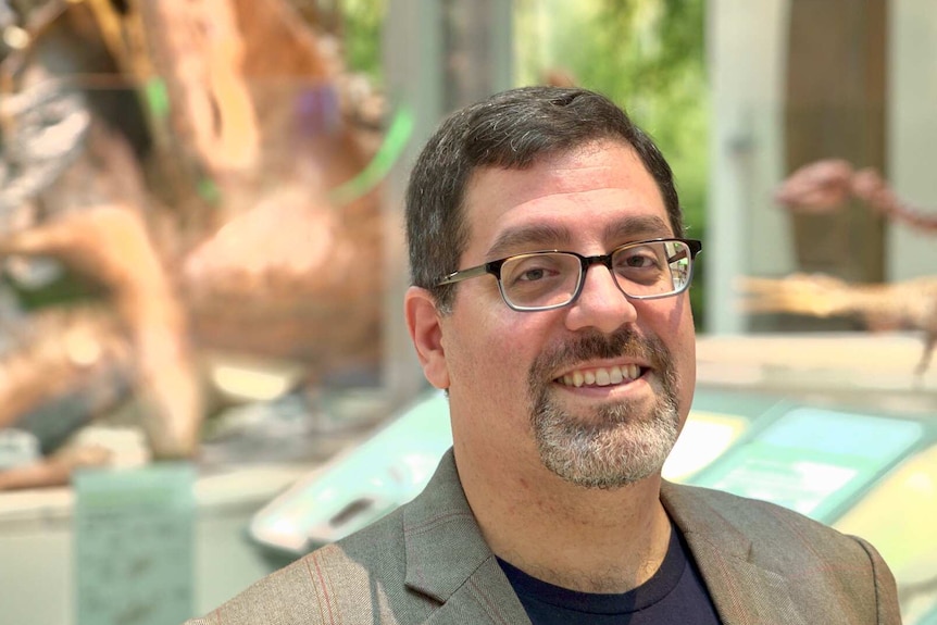A man in glasses smiles while standing in front of dinosaur fossils at a museum