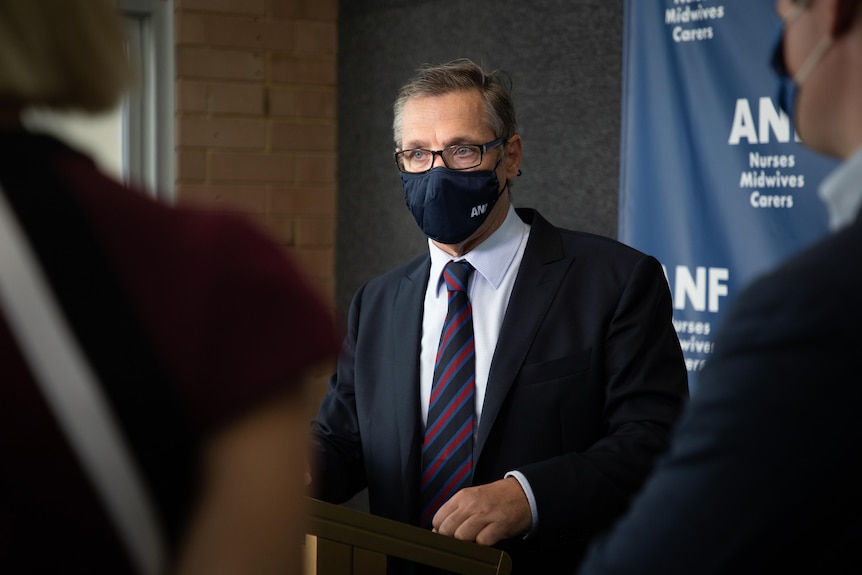 A man in a mask speaks to journalist at a press conference setting
