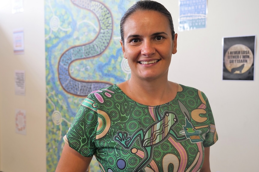 A woman with dark eyes, dark hair, smiles wearing colourful shirt with Aboriginal artwork behind.