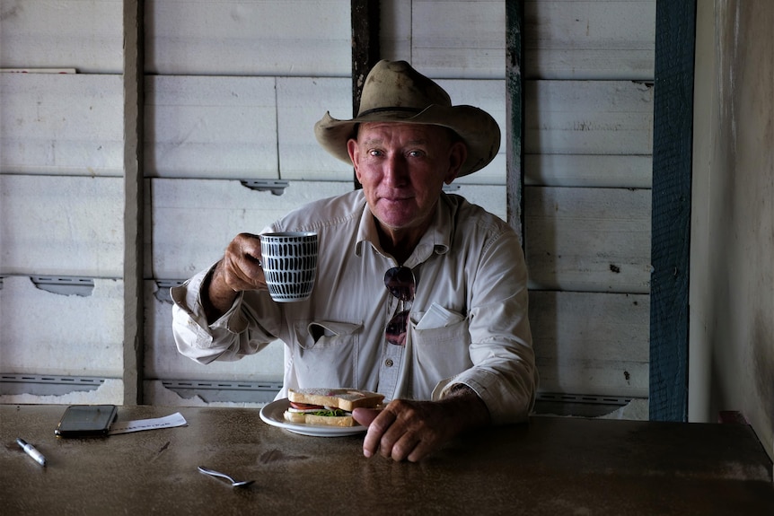A man sits smiling with his mug raised and a sandwich on the table