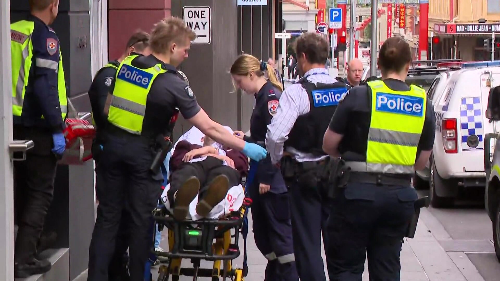A man lies on an ambulance stretcher surrounded by police officers in yellow high vis vests on a city street.