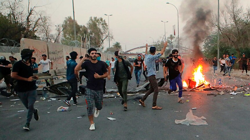 Iraqi protesters run along street past a small fire in Basra