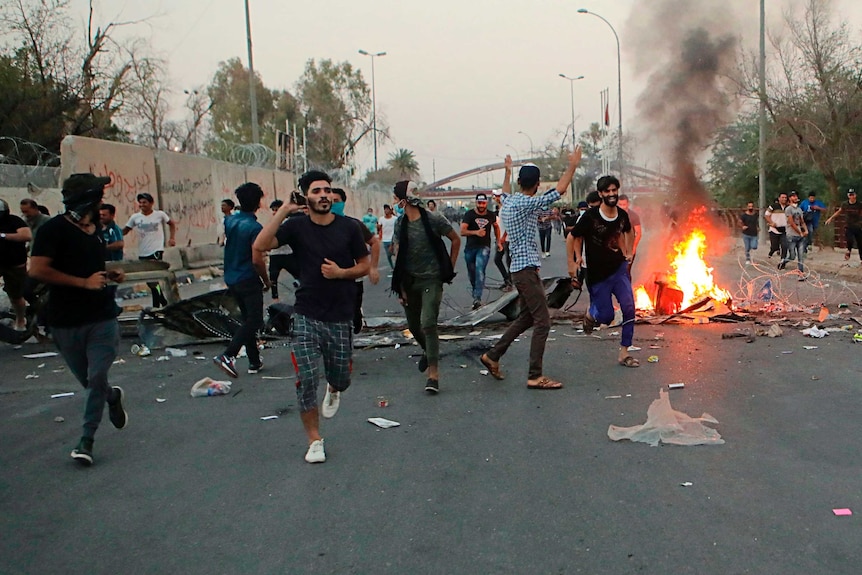 Iraqi protesters run along street past a small fire in Basra