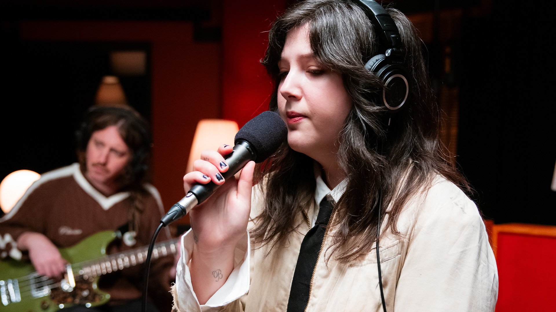 Lucy Dacus wears headphones and sings into a microphone with a bass player behind her