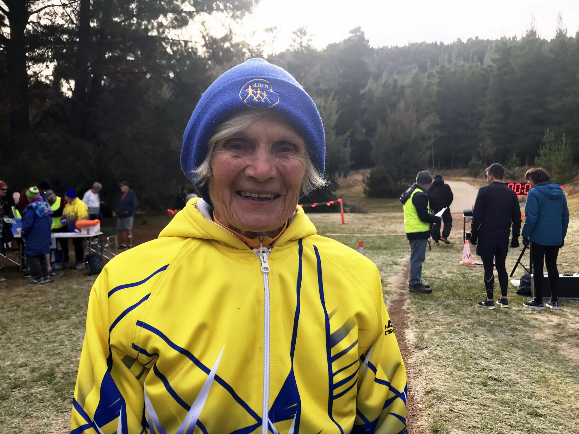An older woman in a bright yellow jackett and blue beanie smiles at the camera.