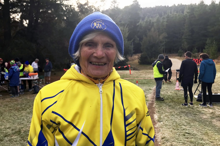An older woman in a bright yellow jackett and blue beanie smiles at the camera.