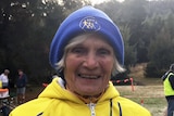An older woman in a bright yellow jackett and blue beanie smiles at the camera.