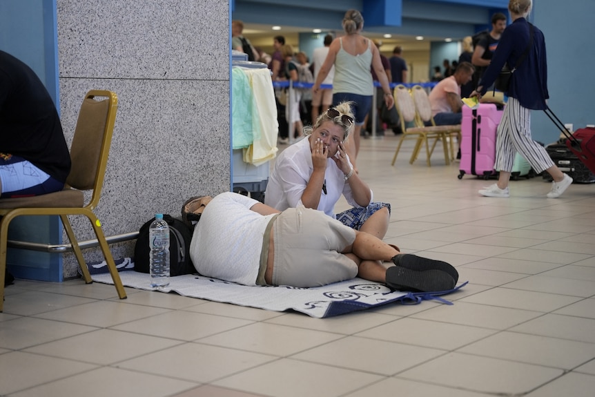 Man lying on airport floor next to woman with blonde ahir looking on 