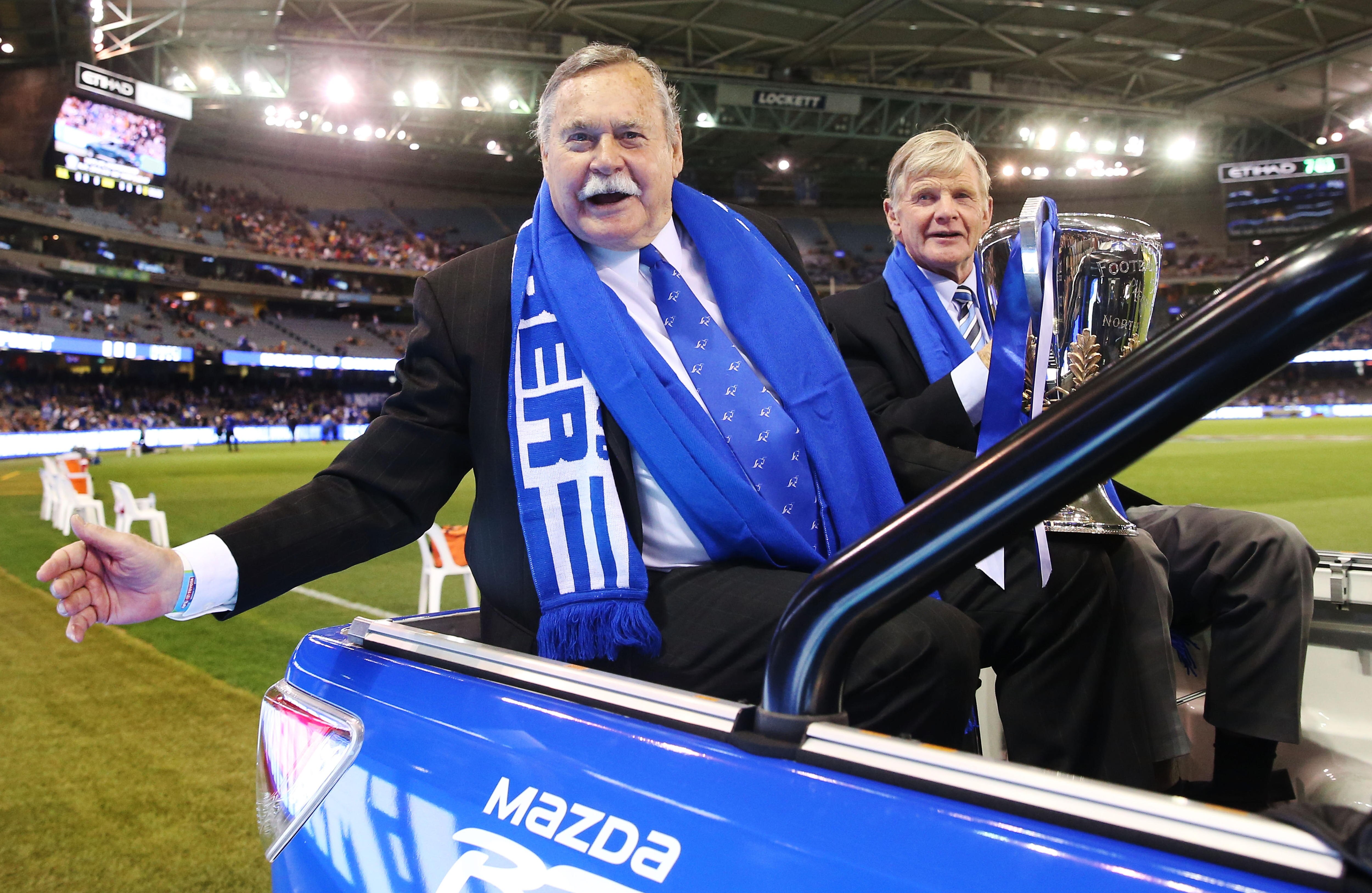 Two older mean in suits and blue scarfs ride a motorcade, carrying an AFL Premiership Cup