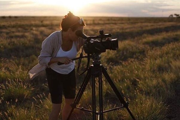 Sunset shot of woman operating a camera on tripod in long grass.