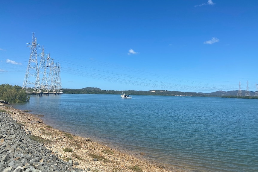 Rocks, power lines and a boat sitting on a body of water, mountains and blue sky behind.