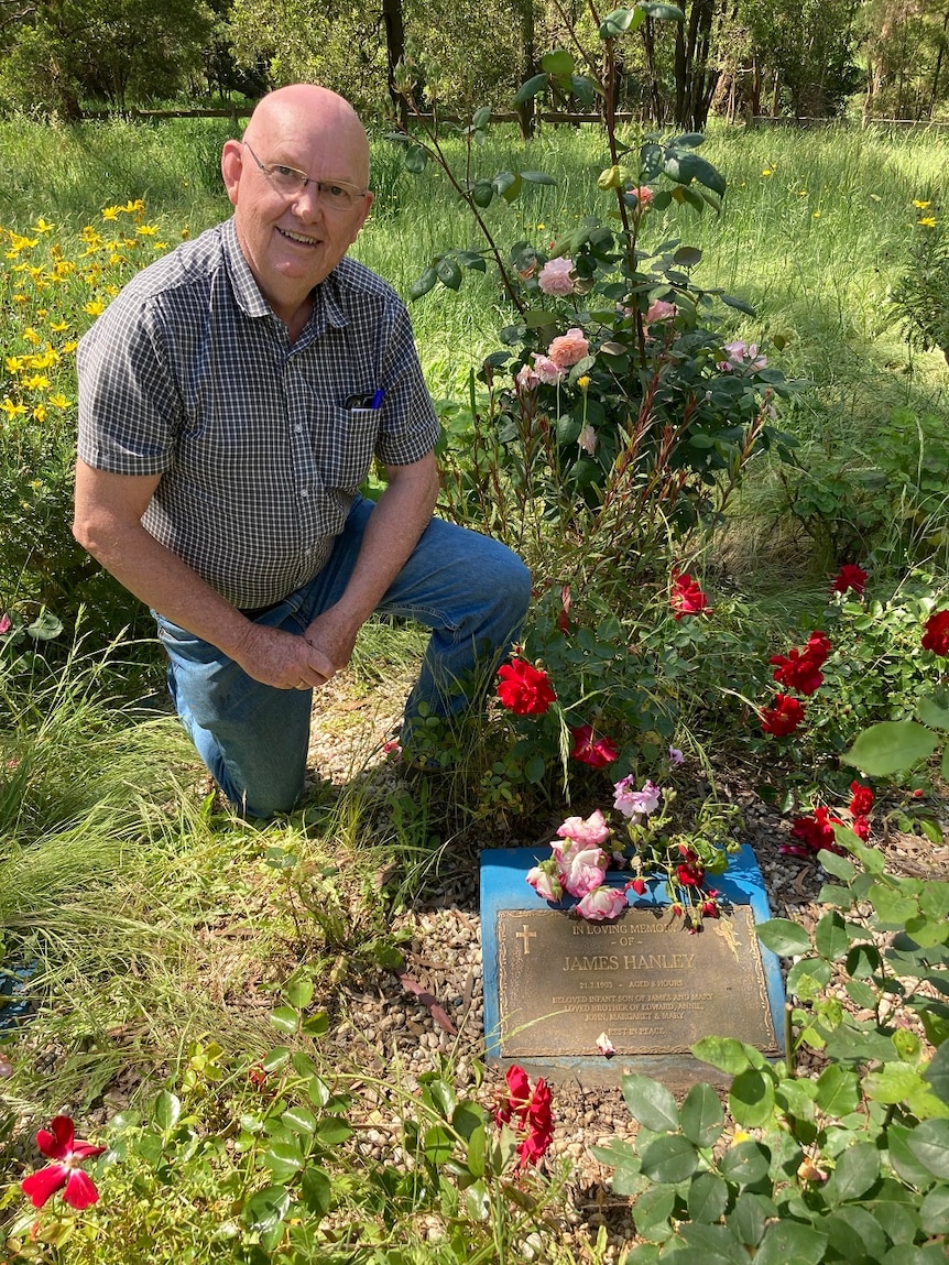 A middle-aged man kneels next to an overgrown grave, with red flowers nearby