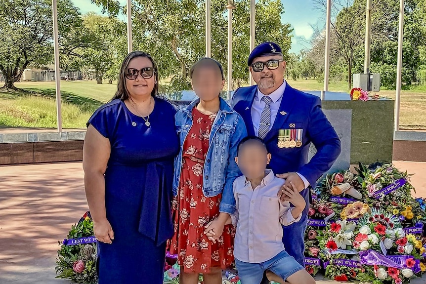 A man in a blue suit with military medals and a beret, standing with a woman and two children with blurred faces, by a memorial
