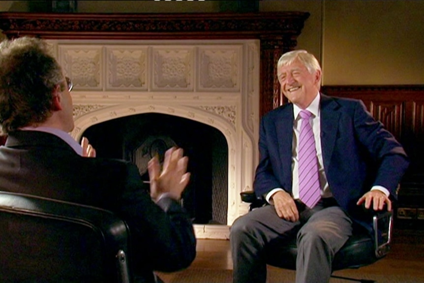 A man sits in a chair with a purple tie while talking to another man.