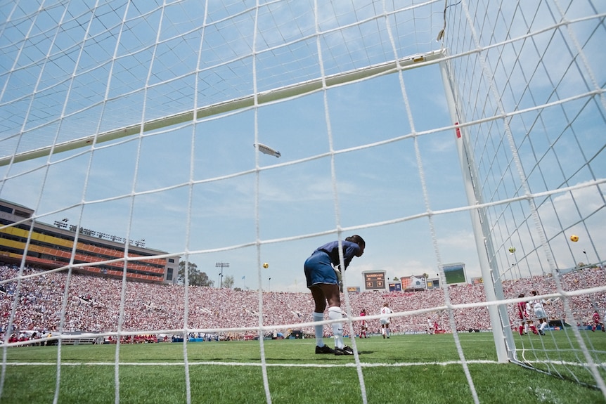 A view through a soccer net shows a goalkeeper, with players and the ball in the background in a packed stadium. 