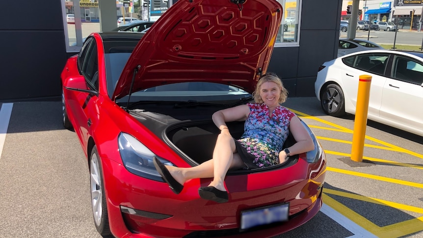 A woman wearing a floral dress sits inside the bonnet of a red car