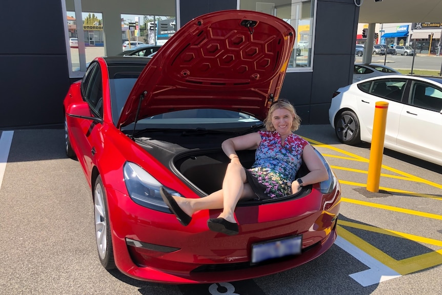 A woman wearing a floral dress sits inside the bonnet of a red car