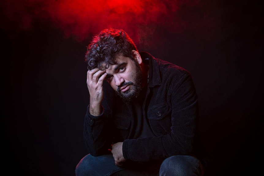 Colour portrait of rapper Philly sitting in front of black background and some red light.