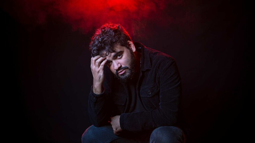 Colour portrait of rapper Philly sitting in front of black background and some red light.