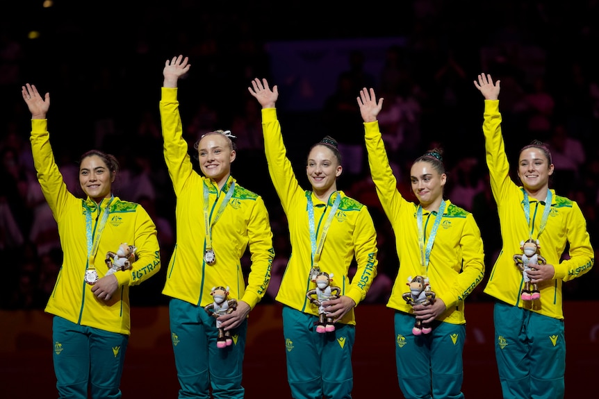 five members of the australian commonwealth games gymnastics team smile and wave on a podium with silver medals on