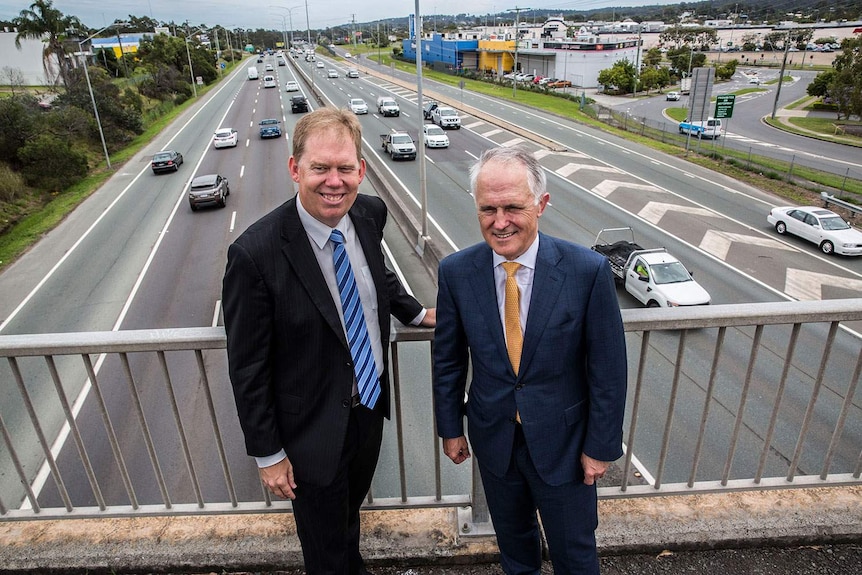 Two men, wearing suits, stand on a highway overpass