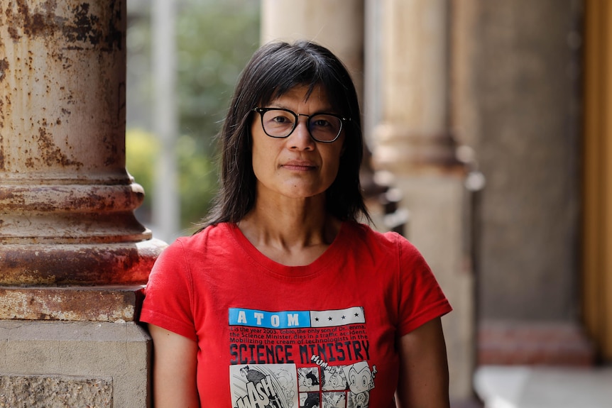 A woman in a bright red t-shirt and glases leans on a pillar.