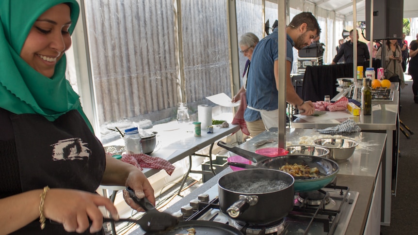 Two people cook at free-standing kitchen benchtops, radio broadcasting equipment and people in background.