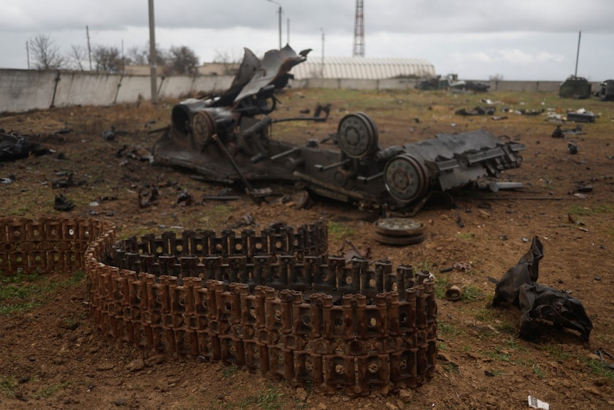 A burned shell of a tank lies upside down on muddy ground, surrounded by miscellanous debris